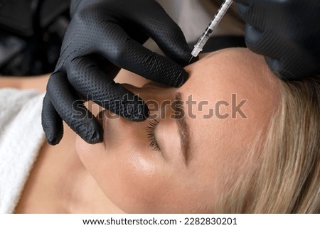 Closeup of Caucasian woman gets botulinum toxin cosmetic injection in forehead. Beauty physician inserts needle of syringe into between the eyebrows, face of female. Cosmetologist, aesthetic medicine.