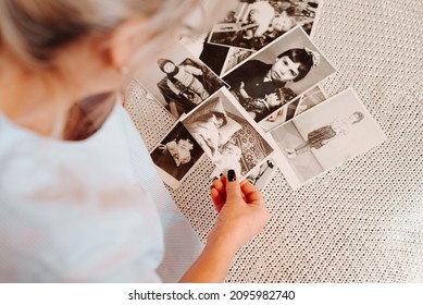 Close-up caucasian woman looking retro photographs of childhood and youth, nostalgic sitting on sofa at home. Back view of female hands holding black and white film photos. Selective focus on pictures