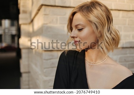 Close-up of caucasian young cute girl in profile against background of blurred light brick wall. Short haired blonde looks calmly down, wearing black blouse with open shoulder.
