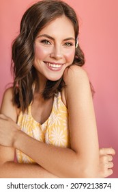 Close-up Of Caucasian Young Girl With Snow-white Smile On Her Face Stands On Pink Background Hugging Herself. Beauty With Wavy Brown Hair, Light Makeup And Yellow T-shirt.