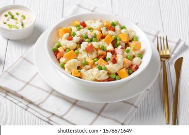 Close-up Of Cauliflower Salad With Crispy Fried Bacon, Green Peas And Cheddar Cheese Cubes In A White Bowl On A Wooden Table With Golden Cutlery, View From Above