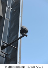 Close-up of a CCTV security camera mounted on the exterior wall of a modern building, captured against a clear blue sky. Concept of surveillance, safety, privacy, monitoring technology, and urban security system.