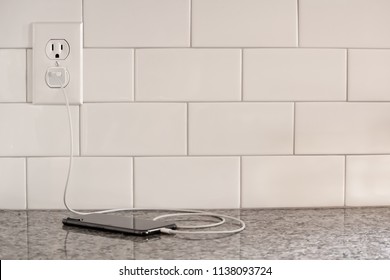 A Closeup Of A Cell Phone Plugged In To A White Subway Tile Wall Outlet Charging On A Granite Countertop, Horizontal Shot, Straight On, Color