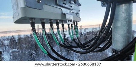 Close-up of cellular base station cables and equipment on a tower above a snowy winter landscape, illustrating telecom infrastructure and wireless network technology.