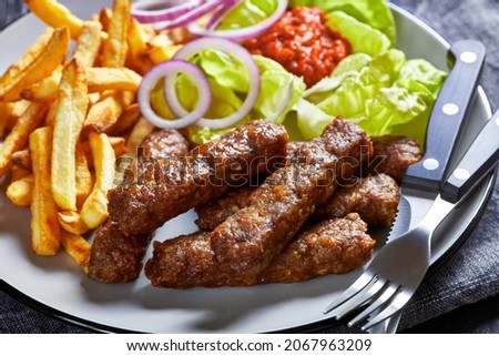 close-up of cevapcici, grilled balkan sausages with potato fries, red onion rings, ajvar and fresh lettuce leaves on a plate on a black wooden table