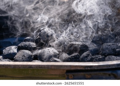 Close-up of charcoal briquettes heating in a grill with white smoke rising. The coals are still mostly black, indicating the early stage of lighting.