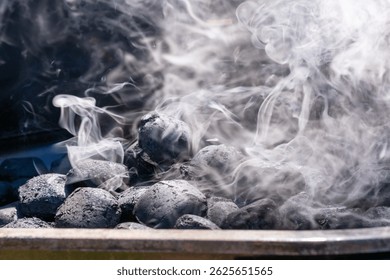 Close-up of charcoal briquettes heating in a grill with white smoke rising. The coals are still mostly black, indicating the early stage of lighting.