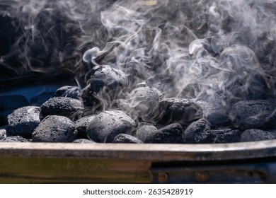 Close-up of charcoal briquettes heating in a grill with white smoke rising. The coals are still mostly black, indicating the early stage of lighting.