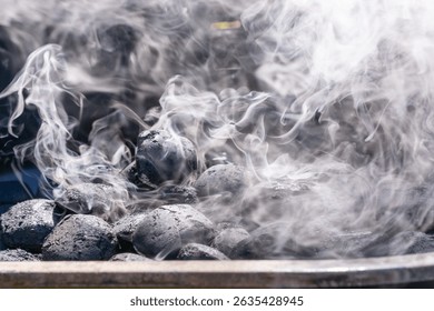Close-up of charcoal briquettes heating in a grill with white smoke rising. The coals are still mostly black, indicating the early stage of lighting.