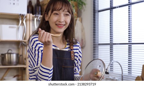 Closeup Charming Asian Female Holding Pot Lid Is Tasting Freshly Made Soup With Wooden Spoon, Feeling Satisfied. Genuine Lifestyle.