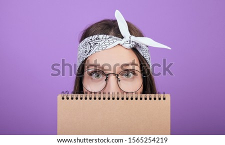 Close-up. A charming green-eyed brown-haired woman in round glasses and a scarf on her head, holds a brown notebook in her hands and hides behind it. Photo on a purple background. Copyspace.