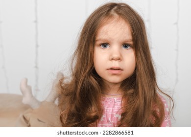 Close-up of a charming little girl with a long loose hair and in a pink pajama, closely, attentively looking at the camera.