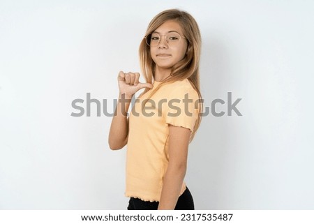 Closeup of cheerful beautiful blonde teen girl wearing orange T-shirt over white studio wall looks joyful, satisfied and confident, points at himself with thumb.