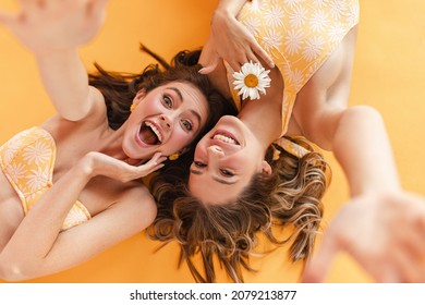 Close-up of cheerful young caucasian female friends fooling around, posing for camera, on yellow background. Smiling broadly, long-haired brown-haired women lie relaxed on floor.