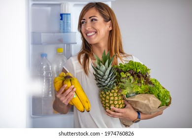 Closeup Of A Cheerful Young Couple Picking Some Fruit And Veggies From The Fridge To Make Some Healthy Breakfast On Sunday Morning. A Side-view Shot Of A Mature Caucasian Woman Taking Something 