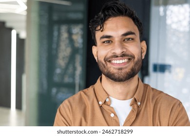 Close-up of a cheerful young man in casual attire, standing confidently in a contemporary office setting. The image exudes professionalism and approachability. - Powered by Shutterstock