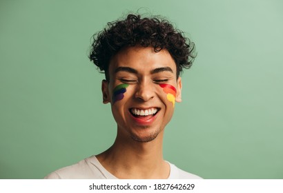 Close-up of a cheerful young man with pride flag painted on face. Gay man with rainbow face paint smiling against pastel green background.