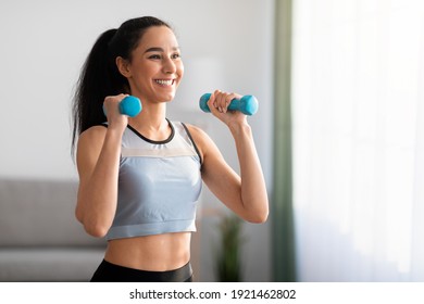 Closeup of cheerful young woman doing dumbbell workout at home, working on arms strength, looking at copy space. Smiling athletic lady lifting blue fitness dumbells up over living room interior - Powered by Shutterstock