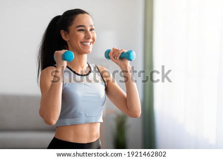 Closeup of cheerful young woman doing dumbbell workout at home, working on arms strength, looking at copy space. Smiling athletic lady lifting blue fitness dumbells up over living room interior