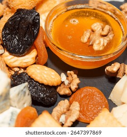Close-up of a cheese platter with honey, dried fruits, crackers, and nuts on a black slate background, representing gourmet appetizer concept