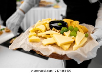 Close-up of a cheese platter with various cheeses, grapes, and nuts on a wooden board. Served by a person in white gloves against a blurred background - Powered by Shutterstock