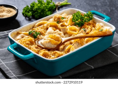 close-up of cheesy and creamy chicken and shells pasta bake in baking dish on dark grey concrete table, horizontal view from above