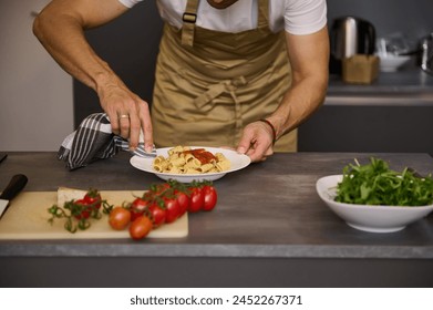 Close-up chef in beige apron, using kitchen towel, removing traces of tomato sauce on the white plate with Italian pasta before serving the dish. Fresh organic ingredients on the kitchen countertop - Powered by Shutterstock