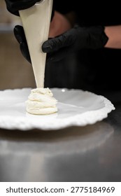 Close-up of a chef in black gloves using a piping bag to swirl white cream or frosting onto a decorative white ceramic plate in a professional kitchen