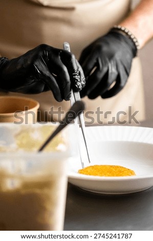 A close-up of a chef in black gloves using kitchen tweezers to carefully place a delicate orange garnish, possibly uni (sea urchin) or citrus zest, onto a white plate during fine dining plating.