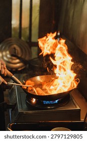 Close-up of chef cooking stir-fry with dramatic fire flame in a frying pan on gas stove.