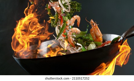 Closeup Of Chef Throwing Prawns From Wok Pan In Fire. Fresh Asian Food Preparation On Dark Background.