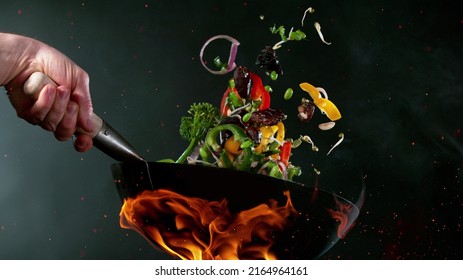 Closeup of chef throwing vegetable mix from wok pan in fire. Fresh asian food preparation on dark background. - Powered by Shutterstock