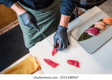 Close-up of chef wearing black gloves slicing raw tuna for sushi. Professional cook preparing fresh fish on cutting board with nigiri sushi in background, showcasing culinary skills and food safety. - Powered by Shutterstock