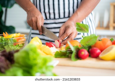 Close-up of chef's hand using stainless steel knife to cut radishes on a wooden cutting board and vegetables such as tomatoes, lettuce and bell peppers prepared on the salad table in the kitchen. - Powered by Shutterstock