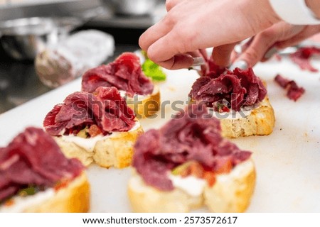 A close-up of a chef's hands adding slices of cured meat onto toasted bread with creamy spread and fresh toppings, showcasing culinary preparation in a professional kitchen.