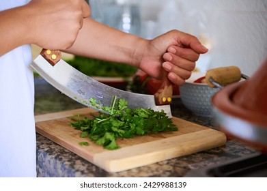 Close-up chef's hands using mezzaluna knife, chopping fresh green herb of parsley on a bamboo cutting board. Preparing healthy food. - Powered by Shutterstock