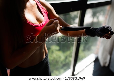 Close-up of the chest of a young woman boxer in a sports top, who ties her arm with black elastic bandages against the background of a panoramic window