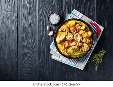 Close-up Of Chicken Curry With Potato, Finely Chopped Red Bell Pepper, Spices In A Black Bowl On A Wooden Table, View From Above, Flatlay