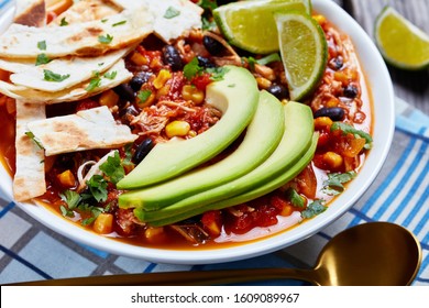 Close-up Of Chicken Taco Soup With Black Bean, Corn Kernels, Topped With Tortilla Strips, Avocado Slices And Lime In A White Bowl On A Rustic Wooden Table, Macro