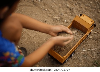 Close-up of a child loading dirt into a yellow toy dump truck with their hands