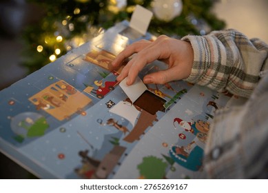 Close-up of a child opening the window of an Advent calendar containing a piece of chocolate, with a Christmas tree adorned with string lights in the background.