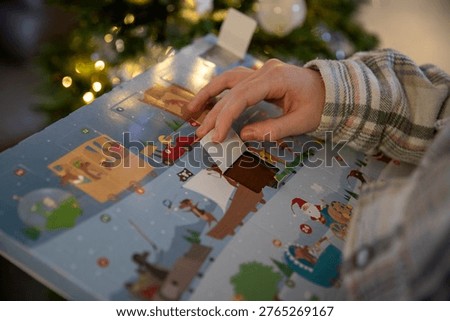 Close-up of a child opening the window of an Advent calendar containing a piece of chocolate, with a Christmas tree adorned with string lights in the background.