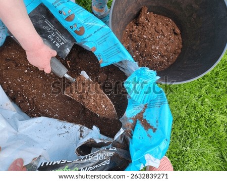 Close-up of a child using a small garden trowel to scoop potting soil from an open bag into a black container on the grass. This image highlights early involvement in gardening and outdoor learning. 