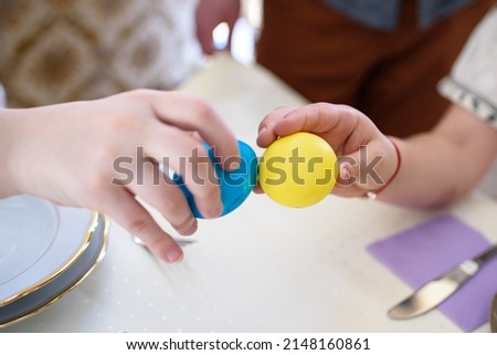Close-up of children's hands holding colorful blue and yellow easter eggs easter eggs. Table, covered with a white cloth, with festive traditional food. Ukrainian Easter eggs. Easter fun, tapping eggs