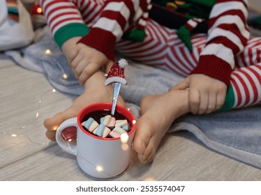 Close-up of a child's bare feet, dressed in Christmas pajamas, holding a cup of cocoa and marshmallows on the floor with his feet. winter weekends and holidays. Lifestyle.