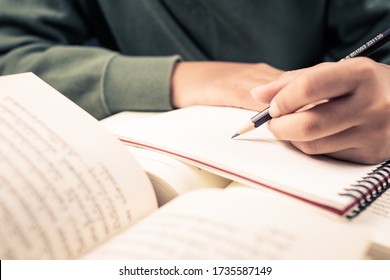 Closeup Child's Hand Going To Write On Notebook With The Opened Novel Book On The Desk, Summarize The Story, Edit A Note After Reading