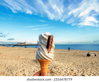 Close-up of a classic British soft whip ice cream in a cone, with a flakey type of chocolate bar pushed into the ice cream held up with a view of sunny Bournemouth beach and pier in soft focus behind - Powered by Shutterstock