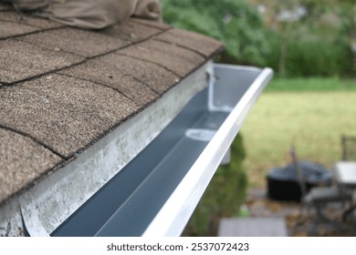 A close-up of a clean, freshly installed house gutter along a roof with brown shingles. The seamless gutter appears free from debris, ready to handle rainwater efficiently.