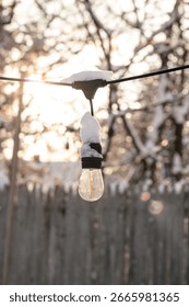 A close-up of a clear outdoor string light bulb covered in fresh snow, glowing softly in the winter sunlight with a blurred wooden fence and trees in the background