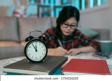 Closeup Of A Clock Against Blurred Background Of A Diligent Japanese Girl Burning Midnight Oil. Asian Female Student Still Up Studying Late At Night